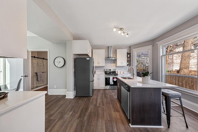 kitchen featuring wall chimney range hood, sink, appliances with stainless steel finishes, white cabinetry, and a barn door