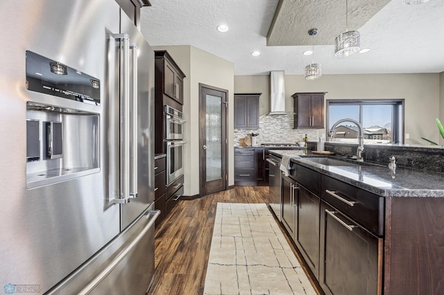 kitchen featuring stainless steel appliances, a sink, wall chimney range hood, dark brown cabinetry, and wood finished floors