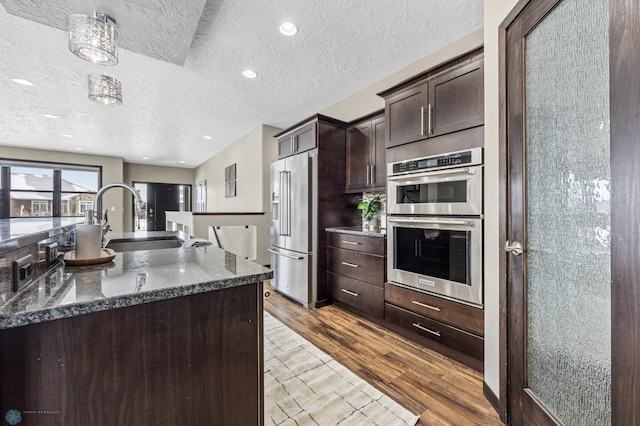 kitchen with appliances with stainless steel finishes, a sink, dark brown cabinets, a textured ceiling, and wood finished floors