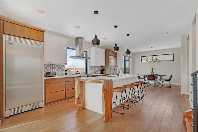 kitchen with white cabinetry, extractor fan, an island with sink, backsplash, and stainless steel built in refrigerator