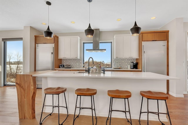 kitchen with island exhaust hood, stainless steel built in fridge, a kitchen island with sink, hanging light fixtures, and white cabinets