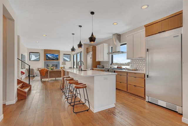 kitchen with built in fridge, exhaust hood, white cabinetry, light hardwood / wood-style floors, and a center island with sink