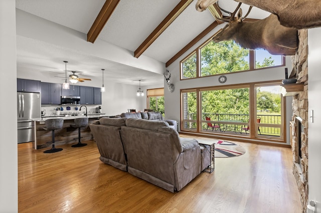 living room featuring a stone fireplace, light wood-type flooring, high vaulted ceiling, beam ceiling, and ceiling fan