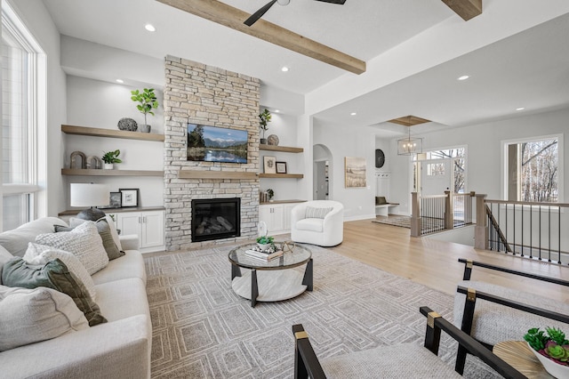 living room with ceiling fan, light hardwood / wood-style flooring, a stone fireplace, and beamed ceiling