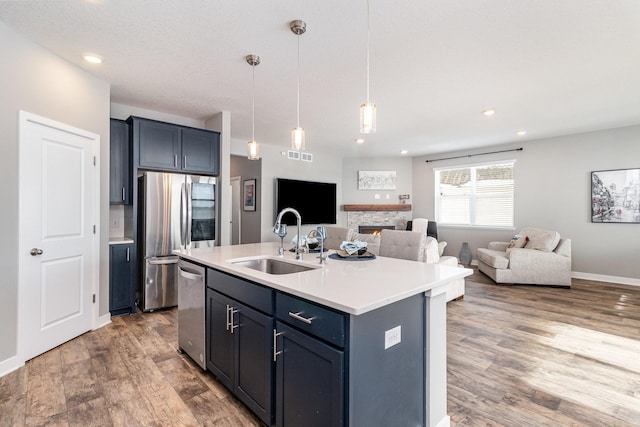 kitchen with pendant lighting, appliances with stainless steel finishes, a stone fireplace, sink, and a center island with sink