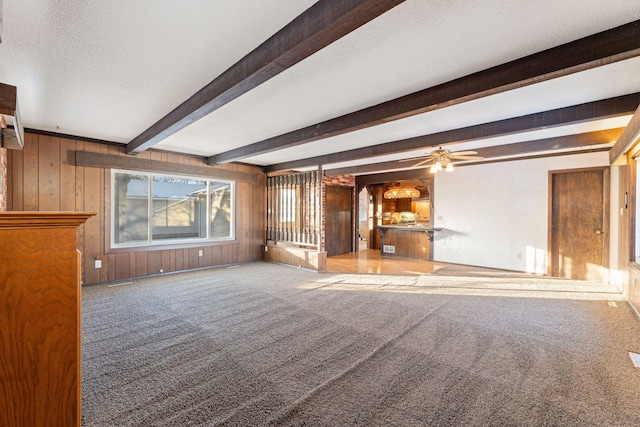 unfurnished living room with ceiling fan, beam ceiling, wooden walls, carpet floors, and a textured ceiling