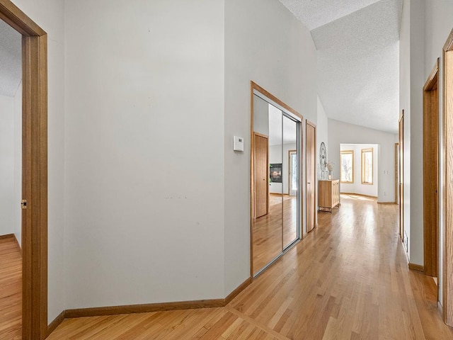 corridor with a textured ceiling, light wood-type flooring, baseboards, and vaulted ceiling