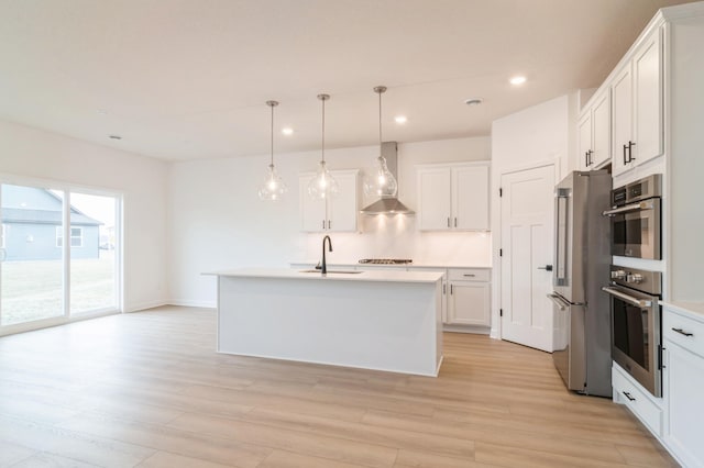 kitchen with white cabinetry, wall chimney range hood, decorative light fixtures, stainless steel appliances, and a center island with sink