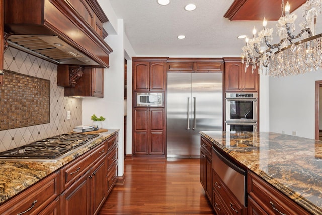 kitchen with dark stone countertops, backsplash, built in appliances, custom range hood, and decorative light fixtures