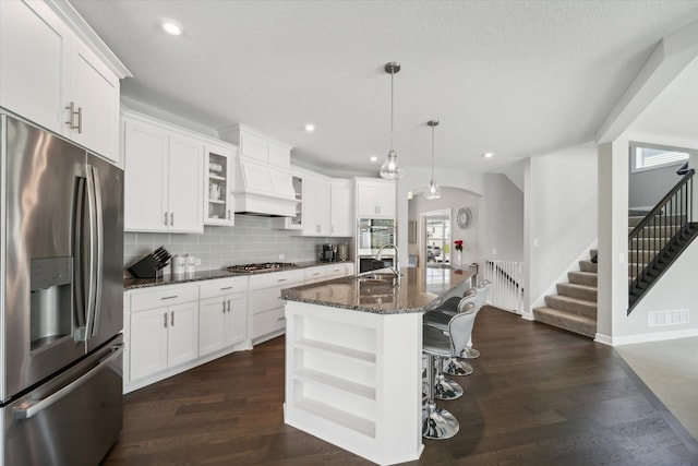 kitchen with an island with sink, appliances with stainless steel finishes, decorative light fixtures, dark stone counters, and white cabinets