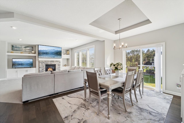 dining space featuring a notable chandelier, a raised ceiling, hardwood / wood-style flooring, a stone fireplace, and built in shelves