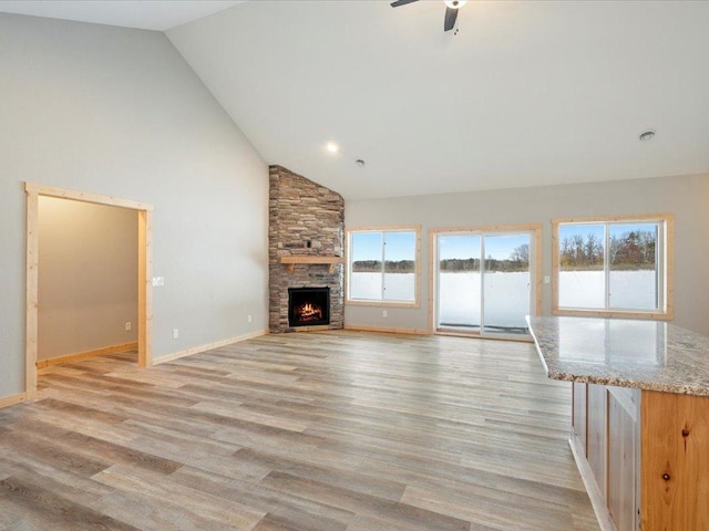 unfurnished living room with high vaulted ceiling, light wood-type flooring, a stone fireplace, and ceiling fan