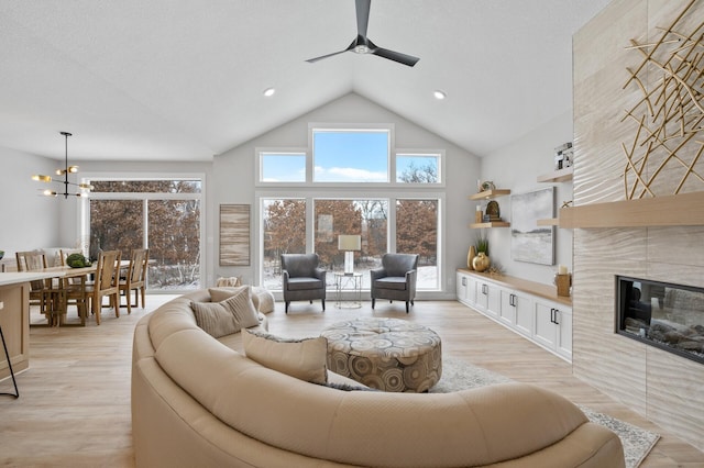 living room with ceiling fan with notable chandelier, a tiled fireplace, a towering ceiling, and light hardwood / wood-style flooring