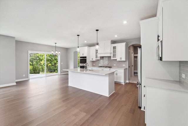 kitchen featuring premium range hood, pendant lighting, decorative backsplash, an island with sink, and white cabinets