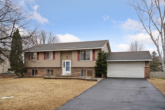 raised ranch featuring driveway, a garage, a shingled roof, fence, and brick siding