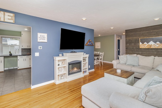 living area featuring an accent wall, a glass covered fireplace, wood walls, light wood-type flooring, and baseboards