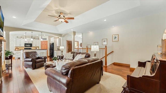 living room featuring arched walkways, a raised ceiling, a ceiling fan, wood finished floors, and ornate columns