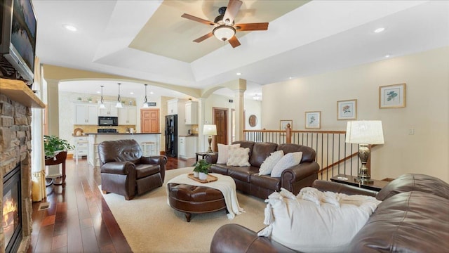 living area featuring decorative columns, arched walkways, ceiling fan, hardwood / wood-style floors, and a stone fireplace