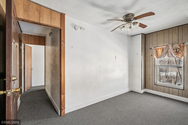 unfurnished room featuring ceiling fan, wooden walls, a textured ceiling, and dark carpet