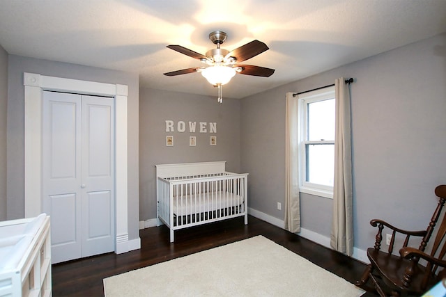 bedroom with a nursery area, dark wood-type flooring, ceiling fan, and a closet