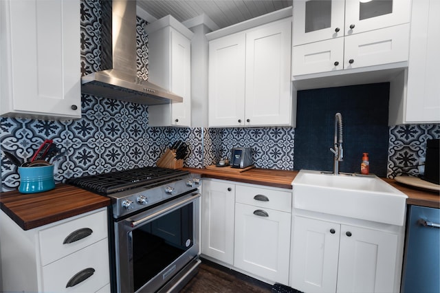 kitchen with butcher block countertops, white cabinets, wall chimney range hood, stainless steel gas range, and sink