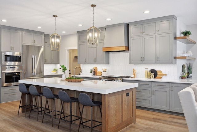 kitchen featuring gray cabinetry, built in appliances, an island with sink, custom exhaust hood, and light wood-type flooring