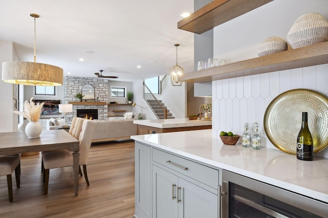 kitchen with ceiling fan, hanging light fixtures, a stone fireplace, beverage cooler, and light wood-type flooring