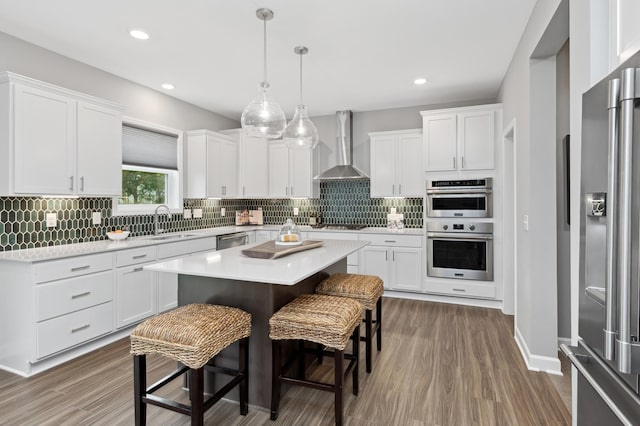 kitchen featuring a kitchen island, white cabinets, hanging light fixtures, stainless steel appliances, and wall chimney exhaust hood