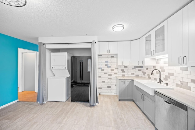 kitchen with black refrigerator, stacked washer / dryer, sink, white cabinets, and stainless steel dishwasher