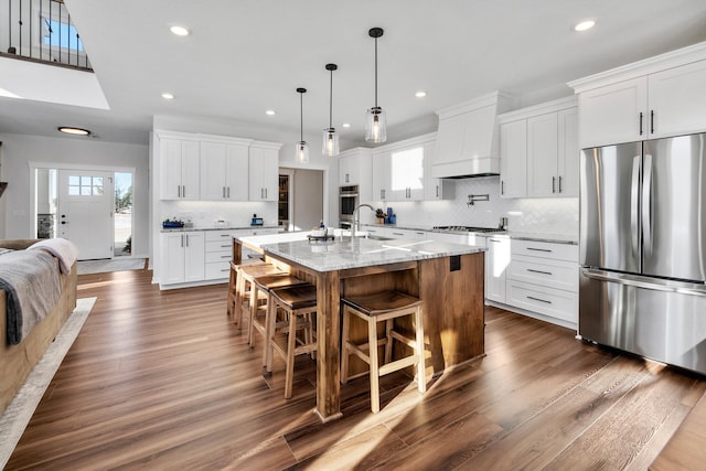 kitchen featuring custom exhaust hood, white cabinetry, decorative light fixtures, appliances with stainless steel finishes, and a kitchen island with sink