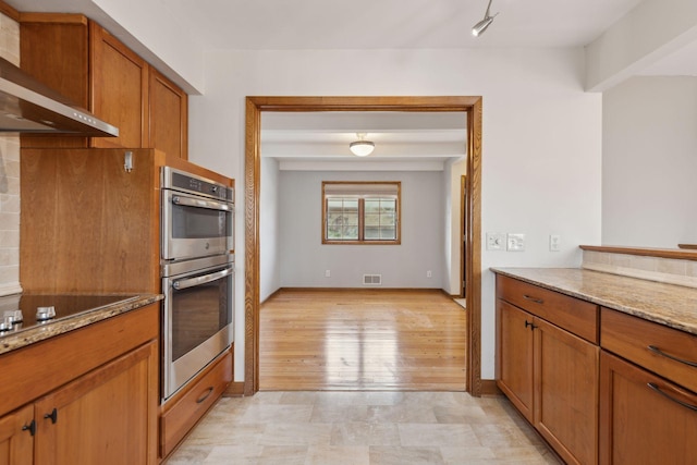 kitchen featuring stainless steel double oven, light stone countertops, and wall chimney range hood