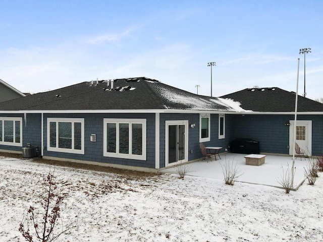 snow covered rear of property with a patio area, central AC unit, and roof with shingles