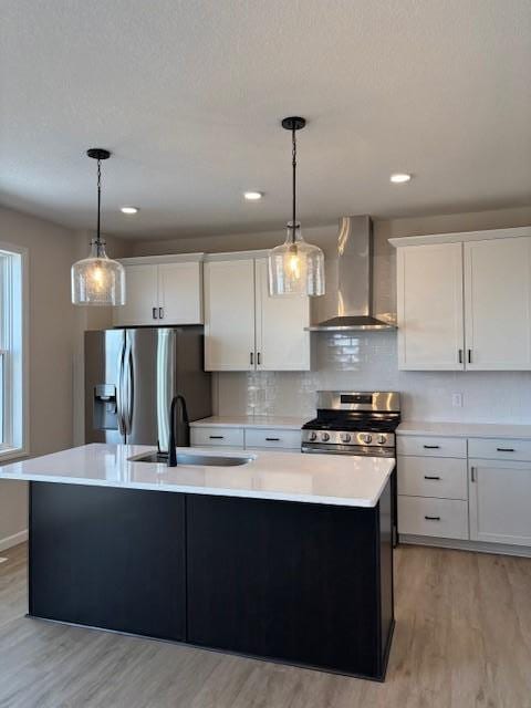 kitchen with an island with sink, stainless steel appliances, light countertops, wall chimney range hood, and a sink