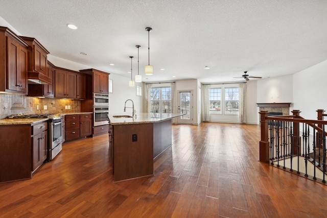 kitchen with pendant lighting, appliances with stainless steel finishes, tasteful backsplash, light stone countertops, and an island with sink