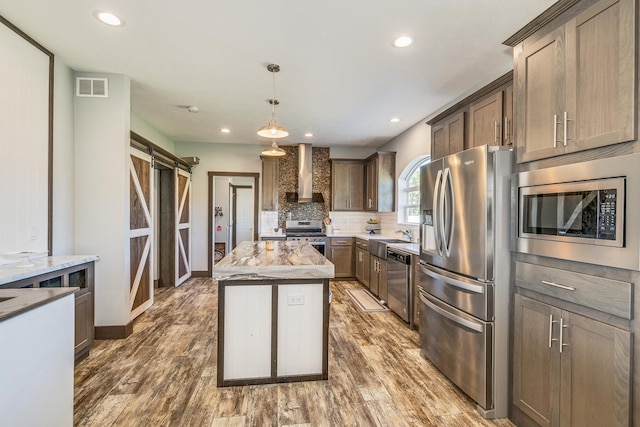 kitchen featuring appliances with stainless steel finishes, decorative light fixtures, a barn door, light stone countertops, and wall chimney exhaust hood