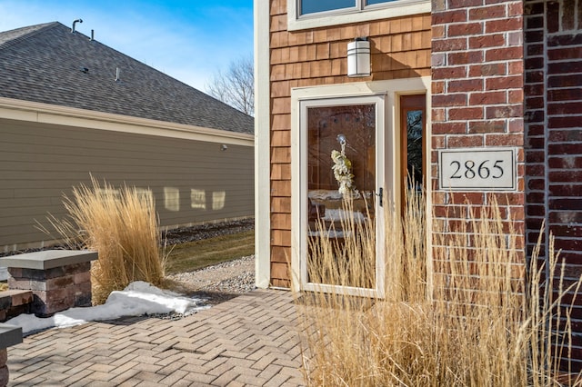 view of exterior entry with a shingled roof and brick siding