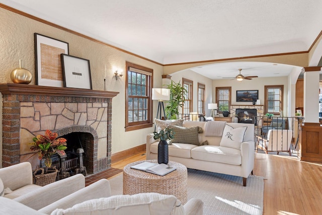 living area with a ceiling fan, light wood-style flooring, arched walkways, a stone fireplace, and crown molding