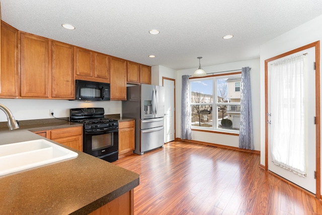 kitchen with brown cabinetry, a sink, a textured ceiling, wood finished floors, and black appliances