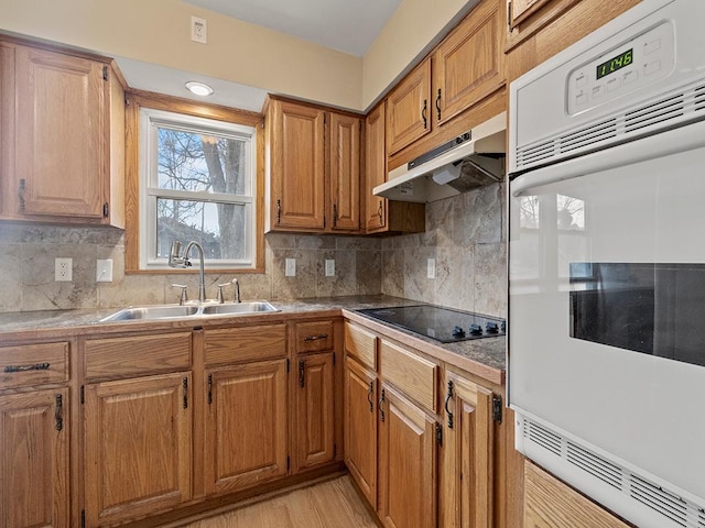 kitchen with tasteful backsplash, brown cabinets, black electric cooktop, under cabinet range hood, and a sink