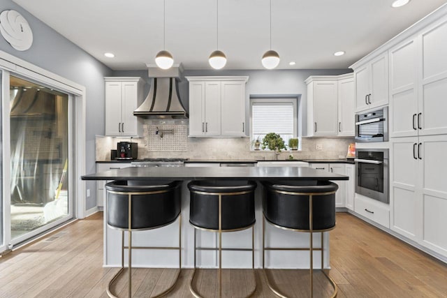 kitchen with wall chimney exhaust hood, stainless steel oven, white cabinets, and decorative light fixtures