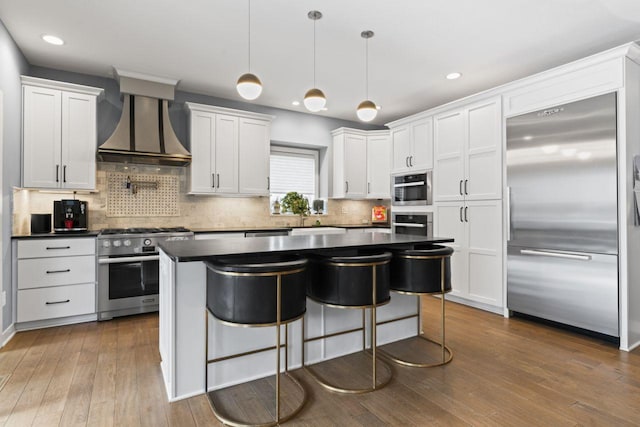 kitchen with wall chimney range hood, appliances with stainless steel finishes, white cabinetry, hanging light fixtures, and dark hardwood / wood-style flooring