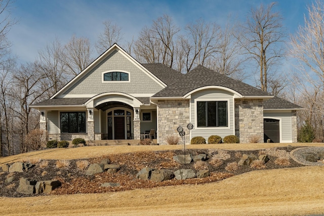 view of front of property featuring an attached garage, covered porch, stone siding, and roof with shingles