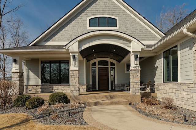 doorway to property featuring stone siding, a porch, and a shingled roof