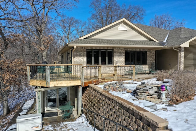 view of snowy exterior with brick siding, roof with shingles, and a wooden deck