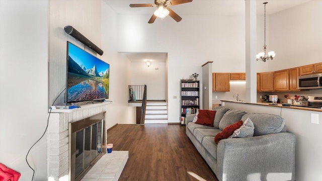 living area with stairway, dark wood-style floors, a fireplace, a towering ceiling, and ceiling fan with notable chandelier