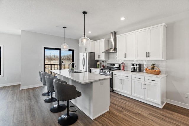 kitchen featuring wall chimney exhaust hood, hanging light fixtures, stainless steel appliances, a kitchen island with sink, and white cabinets