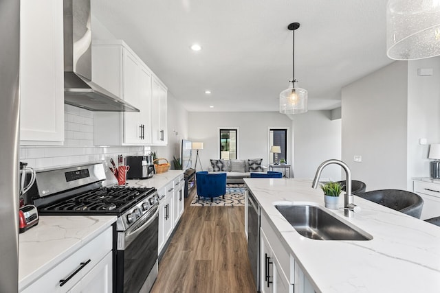 kitchen with appliances with stainless steel finishes, sink, white cabinets, hanging light fixtures, and wall chimney range hood