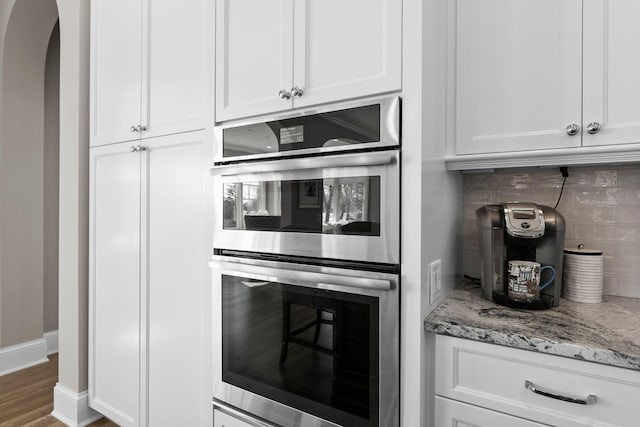 kitchen featuring arched walkways, light stone counters, stainless steel double oven, white cabinets, and tasteful backsplash