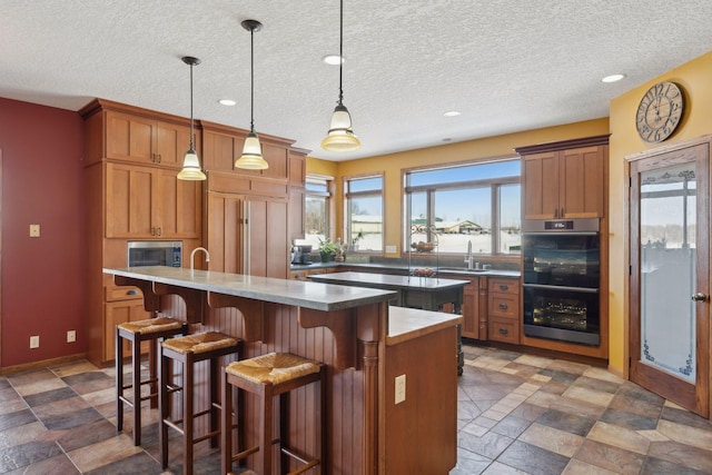 kitchen with hanging light fixtures, brown cabinetry, stainless steel double oven, a kitchen island, and a sink