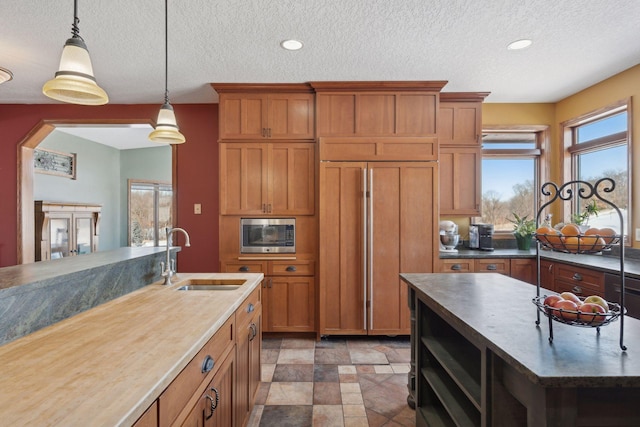 kitchen featuring built in appliances, a sink, brown cabinets, dark countertops, and pendant lighting